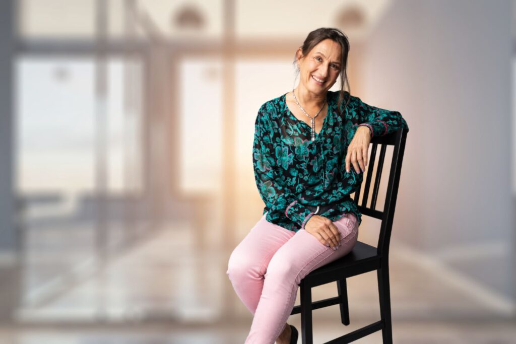 Martina sitzt im Büro auf einem Stuhl, trägt grüngemusterte Bluse und rosa Hose